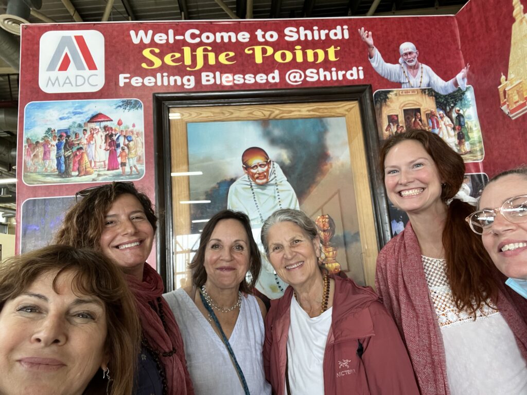 Four women smiling in front of a Shirdi selfie point sign with Sai Baba image.