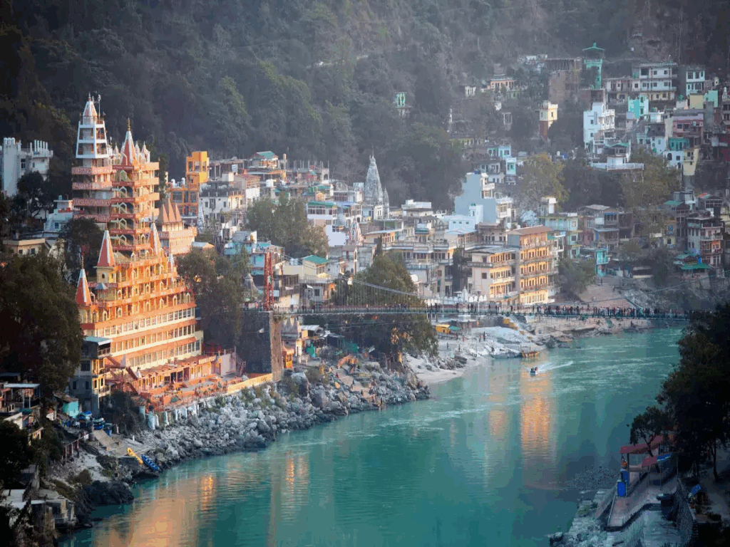 A serene riverside town with temples and buildings nestled against a forested hill.