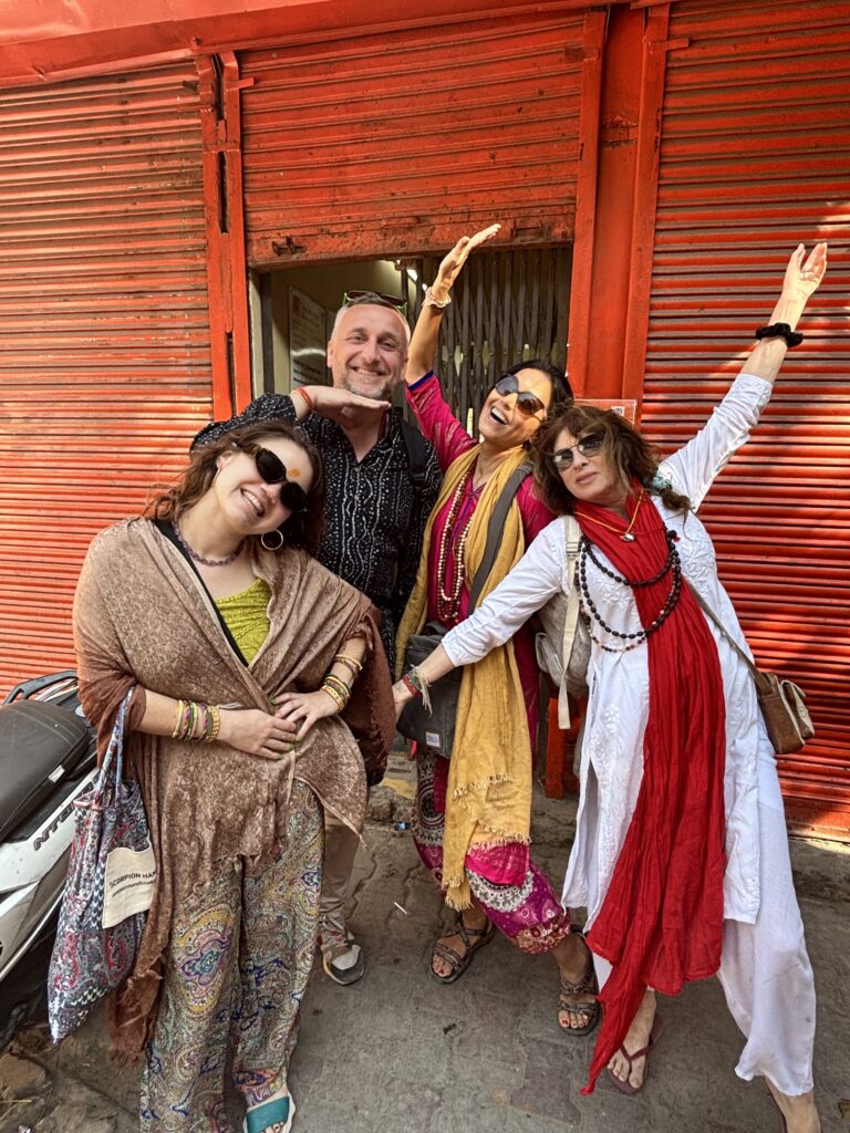 Four women joyfully posing in colorful traditional attire against a red shutter.
